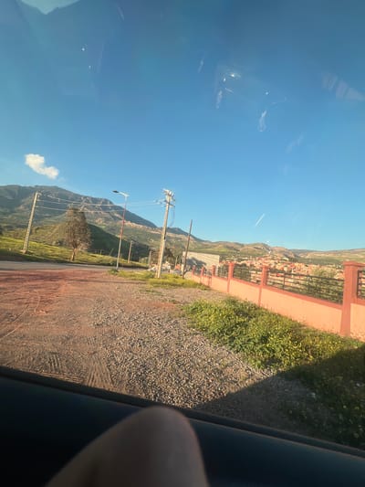 The image, taken from inside a vehicle, shows a road in Ouaouizert, Morocco. The road is unpaved and lined with gravel and vegetation. A pink painted wall with a metal fence runs alongside the road. Beyond the wall, a residential area of the town is visible, nestled in the hills. Power lines and poles are present. The sky is blue with a few clouds. Reflections are visible on the vehicle window. A portion of a person's hand or arm is visible inside the car.
