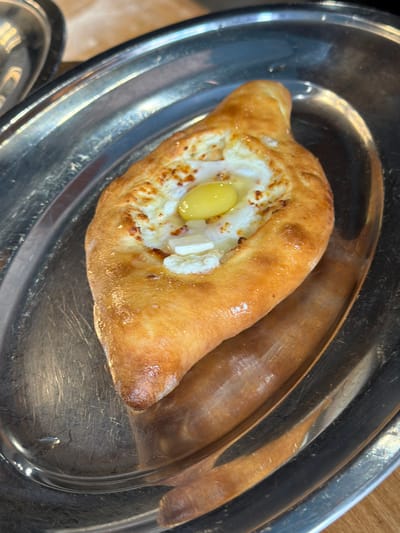This media file captures a close-up, overhead view of a Khachapuri, a traditional Georgian cheese-filled bread, served on a metallic platter. The Khachapuri is boat-shaped with a golden-brown, slightly puffed crust. Inside the bread, a creamy white cheese mixture is visible, topped with a bright yellow egg yolk and some white cheese curds. The egg yolk appears partially cooked, suggesting it was recently added. The platter is oval and made of a reflective metal, showing some scratches and smudges, indicative of frequent use. The background is out of focus, but hints of a wooden surface and a metallic rim of another dish can be discerned. The lighting suggests an indoor setting, possibly a restaurant or home kitchen. There is no visible text in the image. The overall impression is one of a freshly prepared, appealing dish. The image appears to be taken in Maykop, Russia, a location known for its culinary traditions that may include variations of this dish or influences from neighboring cultures.