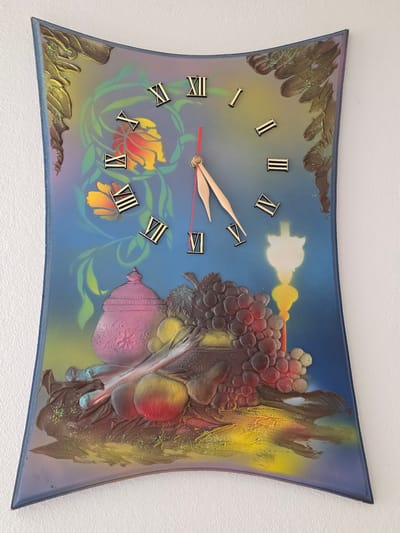 This is an image of a decorative wall clock shaped like a curved rectangle. The clock face is painted with a still life scene featuring a pink pot, grapes, apples, and a candle, all rendered in a somewhat stylized manner. The background transitions from blue to yellow and green, with painted floral elements. Roman numerals mark the hours. The clock hands, one red and one gold, indicate the time. The clock is hanging on a white textured wall. The location of this clock in Herrenberg, Germany, is not determinable from the image.