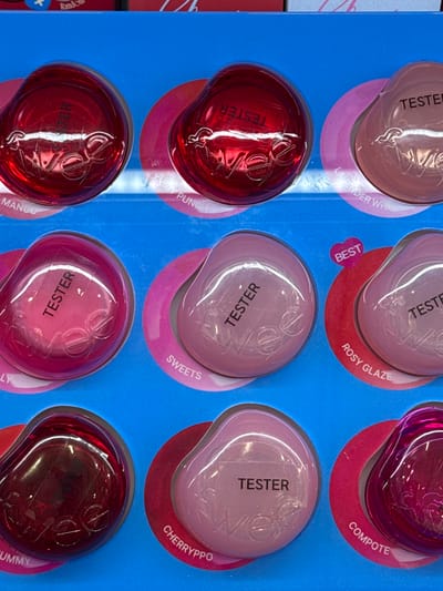 The image displays a retail display of makeup products, specifically lip glosses or balms, arranged in a grid on a blue background. Each product is round and translucent, with a visible brand name "wee" embossed on the surface. Many of the items are labeled "TESTER" with white stickers. The colors of the products range from deep red to various shades of pink and magenta. Some of the products also have color names printed below them, such as "MANGO", "PUNCH", "LAVENDER WHI", "SWEETS", "ROSY GLAZE", "LY", "CHERRYPPO", "UMMY", and "COMPOTE". The overall scene appears to be a point-of-sale display in a retail store. The image itself does not contain any elements that would allow for geographical localization to Nong Pa Khrang, Thailand.