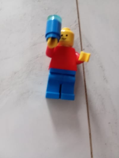 A Lego minifigure, with a yellow head, red torso, and blue legs, stands on a white tiled surface. The minifigure is holding a blue cylindrical object in its right hand, which extends upwards. Its left arm is bent at the elbow, with the hand raised. The background is a white, textured surface, possibly a floor or wall, with faint grey lines suggesting grout or seams. The lighting is neutral, casting minimal shadows. There is no indication of the city of Mũi Né, Vietnam.