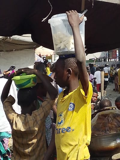 The image depicts a crowded outdoor market scene in Sabon Gari East, Nigeria. A dark fabric canopy partially covers the upper portion of the frame. In the immediate foreground, two young males are prominent. The individual on the right, wearing a yellow short-sleeved shirt with a blue Chelsea FC crest and "3 three." logo, carries a clear rectangular plastic container filled with numerous white, uniformly shaped trapezoidal or rectangular items on his head. His right arm is extended upwards, with his hand resting on the container's lid, holding a small dark object. To his left, a second younger male, wearing a light brown plaid long-sleeved shirt, carries a green and blue lidded plastic bowl on his head, stabilizing it with both hands.

The background is populated with multiple blurred and partially visible individuals, suggesting a busy pedestrian thoroughfare or market area. Structures consistent with market stalls, including white tarpaulins, fabric awnings, and wooden supports, are discernible. In the lower right corner, a large metallic bowl contains dark, possibly fried or cooked food items, partially covered by a clear plastic bag. The scene is brightly lit, indicating daytime conditions.
