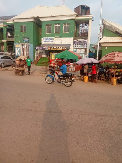 This is a street scene in Kubwa, Nigeria. A blue motorcycle with a rider is traveling on the right side of the road. In the background, a green building with white trim has several windows and a balcony. Signs for "RUFAIDAH YOGHURT & FURA" and "ADAM SHAWARMA" are visible on the building. To the left of the building, two men are standing near a cart with food items. Several market stalls with umbrellas are set up along the sidewalk. A silver car is parked to the far left. The ground is paved with asphalt.