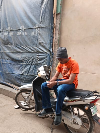 The image displays a young adult male seated on a stationary silver and white scooter-style motorcycle on a concrete surface in Fagge C, Nigeria. The male, positioned facing slightly left, wears a grey knitted beanie, an orange short-sleeved t-shirt with a graphic print, blue jeans, black socks, and black-and-grey Adidas slide sandals. A silver-toned wristwatch is visible on his left wrist. He holds a light-colored mobile phone in both hands, looking downwards at the device. The motorcycle's rear wheel and kickstand are visible, along with a partially obscured green and white Nigerian license plate. The background on the left is dominated by a large blue tarp or canvas, secured horizontally with light-colored ropes, bearing white Chinese characters, including "锦达篷布" (Jin Da Tarp). To the right, a light brown/beige plastered wall features weathered green paint residue near a vertical wooden or metal support structure. The scene is illuminated by natural daylight.