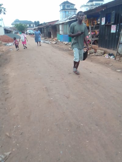 The image depicts a dirt road in Owerri, Nigeria. A man walks towards the viewer, holding a phone and a black bag. A woman and two children with backpacks walk in the opposite direction. Shacks or shops line the side of the road. Trash is visible. A building is in the background.