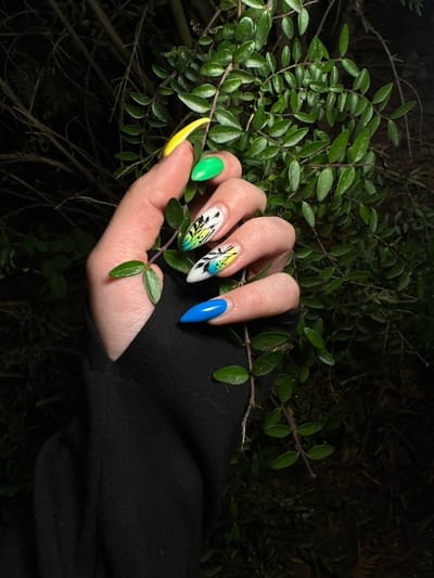 In the city of Dragoman, Bulgaria, a close-up shot captures a person's hand adorned with vibrantly colored, custom-designed acrylic nails. The nails feature a striking combination of bright yellow, lime green, electric blue, and intricate black and green floral patterns on a white base. The hand, with its manicured fingers, is positioned amongst the lush, dark green leaves of a dense shrubbery. The background is dimly lit, with the dark foliage and branches creating a natural, somewhat wild, outdoor setting. The overall lighting suggests it may be dusk or nighttime, as the background is predominantly shadowed and dark. The scene is static, focusing on the aesthetic of the nails against the natural backdrop.
