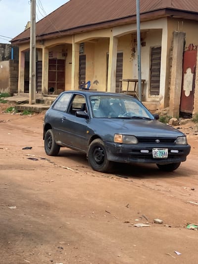 A dark-colored car is parked on a dirt road in front of a multi-story building. The car is a sedan, likely a Toyota, with its license plate visible. The building has several windows and balconies, with some appearing to be boarded up or covered. The ground is unpaved and dusty, with scattered debris. A utility pole with wires runs along the left side of the image. The lighting suggests it could be daytime, possibly overcast. The scene appears to be in a rural or semi-urban area, consistent with a location in Kuson, Nigeria. No people are visible in the frame. The license plate reads "GRE-706AA". There is also some illegible writing on a blue sign above a doorway in the building.