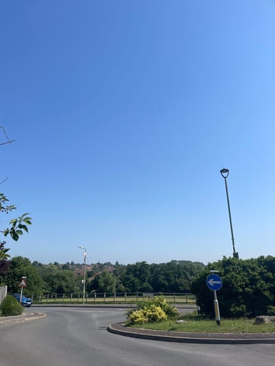 The image depicts a bright, clear sky over a suburban landscape in Maidstone, United Kingdom. The scene is dominated by the expansive blue sky, suggesting a sunny day with good weather. In the foreground, there is a paved road that curves to the right, bordered by a curb and a grassy verge. Adjacent to the road, a wooden fence runs along a slightly elevated area. Beyond the fence, a dense line of trees forms a backdrop, with scattered houses and more trees visible in the distance.
Several man-made objects are present. Two lampposts are visible: one taller, dark-colored lamppost is positioned higher up and to the left, extending horizontally into the sky. Another lamppost, with a curved arm, is closer to the road and appears to have some decorative elements or lighting. A blue traffic sign with a white downward-pointing arrow is mounted on a pole near the taller lamppost. Further down the road, a blue car is parked, and another traffic sign, a warning sign with a red border and a white center, is visible near it. A small, leafy shrub with yellow flowers grows beside the road. In the lower left corner, the branches of a leafy plant with small red berries are visible, adding a touch of nature to the scene.
The composition suggests a daytime setting due to the bright sunlight and the clear sky. The overall mood is serene and peaceful, characteristic of a quiet suburban environment on a pleasant day. No people are visible in the image. The presence of the road, lampposts, and traffic signs indicates an urban or suburban setting, while the extensive greenery points to a location with parks or natural spaces nearby. A faded signpost on the right side of the road has some illegible text.