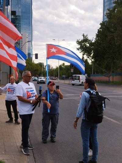 Four individuals are gathered on a city street. Three men hold Cuban and Puerto Rican flags. One man is taking a photo with his phone. Another is wearing a white t-shirt with the words "Queremos libertad" and holding a tripod. The fourth individual, wearing a denim shirt and carrying a backpack, stands facing the group. Tall buildings are visible in the background, and cars drive along the street.
