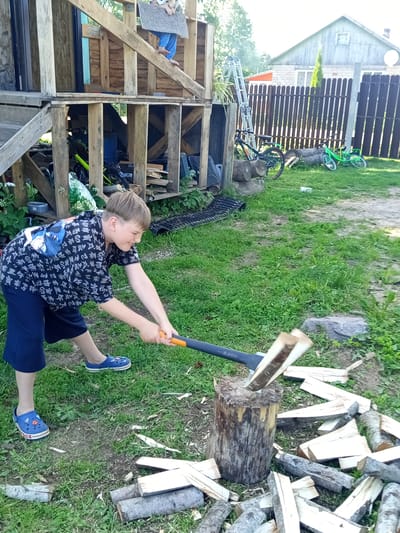 In an outdoor scene in Ostrovtsy, Russia, a young boy is actively engaged in chopping wood. He is wearing a black shirt with a repeating pattern of Asian characters and a blue anime-style graphic on the back, along with navy blue shorts and blue Crocs. The boy is holding a splitting axe with an orange handle and a dark grey head, which is poised to strike a log that is already partially split and resting on a larger chopping block. The ground around the chopping block is covered with a scattering of freshly chopped wood pieces of various sizes, some appearing to be kindling.
The setting appears to be a backyard or a rural area, with a wooden structure, possibly a shed or a multi-level wooden construction, visible to the left of the boy. Behind the wooden structure, a dark brown wooden fence runs horizontally. Two bicycles, one green and another black, are leaning against the fence. The ground is a mix of dirt and green grass, with some patches of dry soil. In the background, a light-colored house with a dark roof is visible. The lighting suggests it is daytime, likely a sunny day given the brightness and the shadows. The overall mood seems to be one of focused activity, possibly a chore or a recreational task. No specific text is legible in the image.