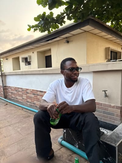 The image displays an outdoor scene in Badore, Nigeria, depicting a single adult male during late afternoon or early evening hours. The individual, dark-skinned with short dark hair, wears a white short-sleeved t-shirt, black trousers, and black-framed glasses. He is seated, oriented slightly to the right of the frame, with a slight smile, and holds a green plastic beverage bottle with both hands, appearing to interact with its cap. Another identical green bottle rests on the reddish-brown tiled ground near his feet, adjacent to a light blue pipe that runs horizontally across the foreground. The male is positioned next to a dark, marble-patterned structure. Behind him, a white wall with a lower section of brown brick patterning features a brass water spigot. In the background, a light-colored building with a dark roof overhang is visible, along with several recessed rectangular window openings. Green tree foliage occupies the upper portion of the frame against a diffused, light-colored sky.
