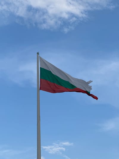 A national flag of Bulgaria is depicted on a flagpole against a backdrop of a bright blue sky with scattered white clouds. The flag is horizontally divided into three equal bands of white, green, and red from top to bottom. The flag is slightly rippled by the wind, with some fraying visible at the edges. The flagpole is tall and appears to be made of metal, positioned to the left of the frame. In the distance, the characteristic red-tiled roofs of houses in the city of Smolyan, Bulgaria, are visible. The overall scene is a clear, outdoor shot on what appears to be a sunny day.
