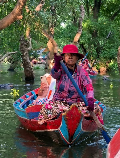 In a narrow waterway surrounded by dense, leafy trees, a woman in a red hat and patterned shirt rows a small, vibrantly painted wooden boat. She is seated at the stern, holding a long oar with both hands, her gaze directed forward. A second person, mostly obscured, is seated in the front of the boat, holding a smartphone. The water is a dark green, reflecting the surrounding foliage. The scene appears to be a canal or river in a rural area, potentially in the Tonlé Sap region near Siem Reap, Cambodia, given the style of the boat and the lush, tropical vegetation. The lighting suggests it is late afternoon or early morning, with dappled sunlight filtering through the trees. The boat itself is colorful, with red, blue, and yellow paint visible.