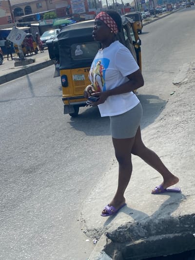 The image captures a female subject standing on a concrete curb next to an asphalt road in Badore, Nigeria, under clear daylight conditions. The subject is a dark-skinned individual wearing a red and white patterned headband, a white short-sleeved t-shirt with a colorful graphic print on the front, and light grey form-fitting shorts. Her feet are clad in purple open-toed slide sandals. She holds a dark, rectangular object, possibly a mobile phone, in her left hand, positioned in front of her. Her head is turned slightly to her right, and her gaze is directed off-frame. Directly behind her, on the asphalt, is a yellow Keke Napep (auto-rickshaw) displaying "TVC" and the license plate "LAGOS GGE-871LC" on its rear. Further down the road, other vehicles and pedestrians are visible. Buildings, appearing to be commercial or residential structures, line the background. The concrete curb shows signs of wear and minor debris. Defined shadows are cast on the ground, consistent with direct sunlight.