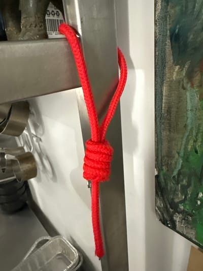 The image shows a red rope tied in a knot around a metal object. The knot is tightly wound, and the rope extends away from it on both sides. In the background, there is a white surface and a painting or print with green and grey tones. There are also other metal objects partially visible below the rope and white surface. The image does not provide any information about the location being Новоглаголево, Russia, or any people, subjects, or actions.