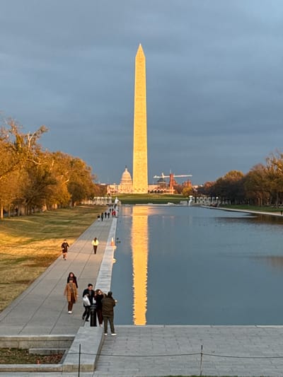 The Washington Monument dominates the frame under a clouded sky. To its right, the U.S. Capitol Building and surrounding structures are visible. A reflecting pool stretches out, mirroring the monument and buildings, with sunlight causing a bright reflection. People walk along a concrete path beside the pool, adding scale to the scene. Trees line the periphery of the area, suggesting a park-like setting.
