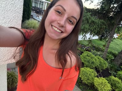 A young woman with long brown hair is taking a selfie outdoors in Bulgarovo, Bulgaria. She is wearing an orange mesh tank top and smiling at the camera. Behind her, a white textured wall is visible on the left, and a lush green garden with various shrubs and trees fills the background. In the distance, a modern house with large windows and outdoor furniture can be seen, suggesting a residential or recreational area. The lighting indicates it is likely daytime, possibly late afternoon or early evening, with soft natural light.