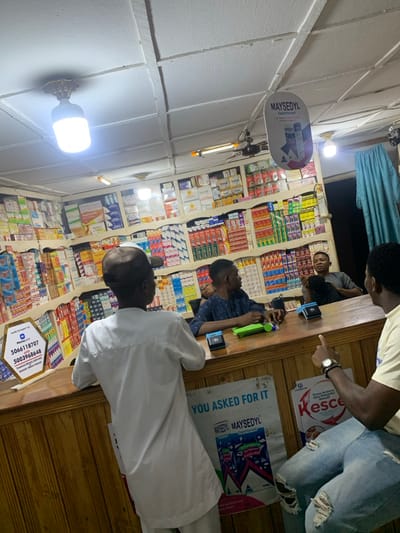 Inside a small store, four people are seen near a counter filled with merchandise. Two are behind the counter, one in a light shirt and ripped jeans pointing, and another in a blue shirt playing with a toy. A third person is partially visible behind the counter. In front of the counter, a person in a white robe and cap is waiting. The shelves behind the counter are stocked with various products, and advertisements for "Mayseydyl" are visible. A hexagon sign with a phone number rests on the counter. The lighting is provided by fluorescent bulbs on the ceiling.