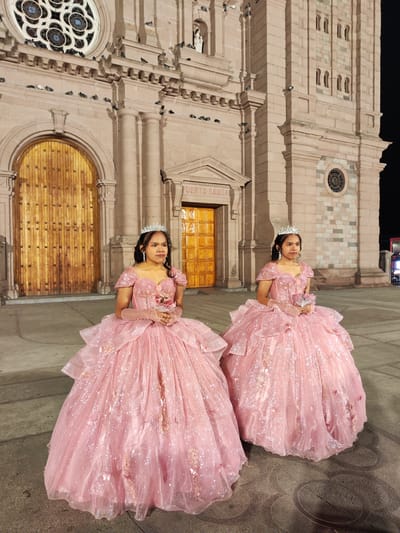 Two girls in pink ball gowns stand on a paved street in front of the Cathedral of Tacna, Peru, at night. Each wears a silver tiara, pink gloves, and holds a bouquet. The cathedral is constructed of light-brown stone. Pigeons perch on ledges and the facade. The arched main entrance and a smaller entrance marked "PUERTA SANTA" are visible. A round window is on the right facade. A modern street light is visible at the right edge.