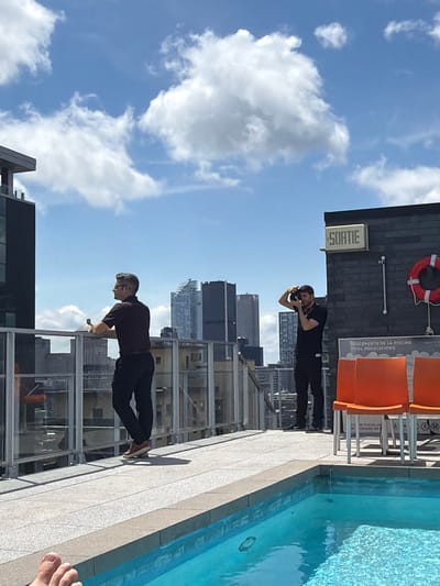 The image depicts a rooftop scene in Montreal, Canada, featuring two men and a swimming pool. On the left, a man wearing glasses, a dark short-sleeved shirt, and dark pants stands near a glass railing, looking to his left. On the right, another man in a black shirt and black pants is taking a photograph with a camera. He is positioned near a brick wall that has a sign with the word "SORTIE" in English, and a red life preserver. In the foreground, a swimming pool with bright blue water is visible, with a person's feet partially in the frame on the lower left. The background displays a cityscape with tall buildings and a cloudy blue sky. The setting appears to be a hotel or residential building's rooftop amenities.