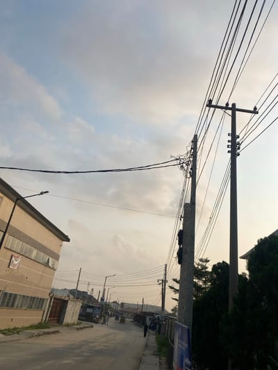 An outdoor scene depicts a street view dominated by utility poles and electrical wires against a cloudy sky. The poles are gray and extend upwards, supporting a network of tangled wires. Buildings line the right side of the street, and a beige structure appears on the left. The street stretches into the distance.