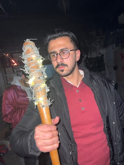 A man with glasses and a mustache holds a baseball bat wrapped in barbed wire and fairy lights. He wears a red sweater under a black puffer jacket. The image is dimly lit, possibly at night. A small fire burns in the background near boxes and other indistinct objects. The man's expression is neutral as he faces the camera.