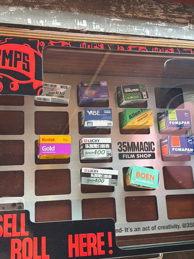 This image depicts a close-up, slightly angled view of a film display case within an establishment in Tibubeneng, Indonesia, identified as "35MMAGIC FILM SHOP." The display case features a grid-patterned, silver-colored metal panel with rectangular cutouts, revealing a dark brown, wood-grain surface beneath where various film boxes are placed. The entire display is observed through a clear, reflective surface, likely glass or acrylic, on which a partial reflection of a hand's thumb is visible on the right.

The film boxes are arranged in rows. Brands include "VIBE" (100, 400 ISO), "WOLFEN UN 54" (Black and White Film 135-36), "Alien Film 5219/500T" (Color Negative Film), "FOMAPAN" (ACTION 400/36, CREATIVE 200/36), "Kodak Gold" (Color Negative Film 135/200), "LUCKY SHD100" (Black and White Film 120), "LUCKY SHD400" (Black & White Film 135-36 and 120), and "BOEN JOGJAKARTA."

Signage on the display includes the central text "35MMAGIC FILM SHOP" in black on a silver background. A black panel at the bottom left reads "SELL ROLL HERE!" in red capital letters. To its right, partial text in black states "nd- It's an act of creativity. @35m". The top left corner features a red stylized "LAMPS" logo or branding on a black background with swirling red accents. The top edge of the frame reveals a distressed wall or surface with white paint flakes on a darker, possibly grey, background. No individuals are directly visible within the frame, apart from the reflection of the person holding the camera.
