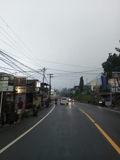 In the city of Dawathang_Dorjibi_ Kashingtsawa, Bhutan, an outdoor street scene is depicted under an overcast, bright gray sky, with atmospheric mist or fog obscuring distant hills. The asphalt road, wet from precipitation, extends towards the background, marked by a solid white line on the left and a broken yellow centerline. On the left side of the road, a male individual wearing a red long-sleeved shirt, light-colored shorts, and a cap stands barefoot on a sidewalk adjacent to a row of commercial buildings. These structures display various signs, including "Canon Digital Copier," "Beauty Make Up," and "ATM." A dense network of utility wires spans overhead, connected to multiple utility poles along the street. Several motorcycles are parked on the sidewalk along the left side. In the mid-ground, vehicular traffic is present on the wet road; a white compact car with illuminated headlights is observed traveling away from the viewer, preceded by a motorcycle. Additional vehicles with headlights on are visible further down the road. The right side of the street features an elevated bank with more commercial signage, including partial views of "RESTORAN" and "VILLA APAYA," and a tall, slender evergreen tree. Several motorcycles are also parked on the shoulder along the right side. The overall lighting suggests either dusk or a heavily overcast day.
