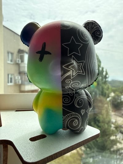 A figure of a stylized bear, half rainbow-colored with a black cross on its forehead and half black with white decorative stars and swirls, stands on a light-colored wooden shelf. The bear is positioned in front of a window, through which buildings and trees are visible. The sky is overcast. The scene is likely indoors, looking out a window, with the figure placed on a shelf. There is no indication of the specific location within Kharkiv, Ukraine, beyond what can be inferred from the general urban and natural elements visible through the window.