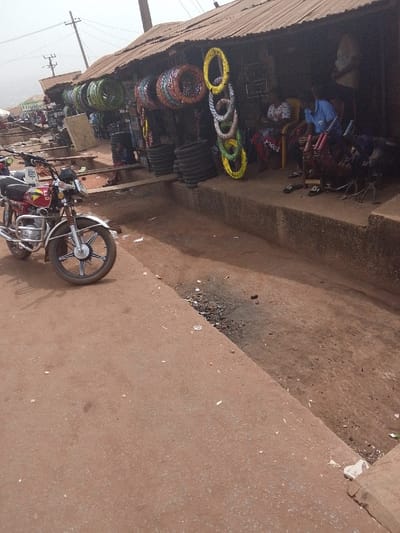 This image depicts a street scene in Ankpa, Nigeria, characterized by commercial activity and unpaved surfaces. In the immediate foreground, a reddish-brown dirt or gravel path occupies the left side, where a red and chrome motorcycle is parked facing the right. The motorcycle features a black front tire with a silver rim and black spokes. To the right of the path, a concrete drainage channel or gutter runs parallel to a row of open-fronted shops. This channel contains dark debris and light-colored refuse.
The primary shop visible on the right features a corrugated metal roof supported by dark posts. The facade of this shop is adorned with various brightly colored motorcycle tires, including green, yellow, orange, and silver, hanging as merchandise. Stacks of black tires are also visible on the ground level within the shop's open front. Inside, two individuals are seated. One person, wearing a patterned dress, sits on a yellow plastic chair. Another person, dressed in a blue shirt, sits nearby, appearing to handle or inspect various motorcycle components, including what resembles handlebars. Black sandals are placed on the ground in front of these individuals. A wooden plank serves as a makeshift bridge over the drainage channel, providing access to the shop.
Further down the street, additional shops with similar structures are visible, along with utility poles and overhead wires against a hazy sky. A distant hill or elevated terrain is faintly visible on the far left horizon. The overall environment suggests an active, utilitarian commercial area with basic infrastructure.