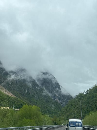 The image depicts a view from a vehicle traveling on a roadway in Monastyr', Russia. A mountain with dense green foliage is partially obscured by fog and low-hanging clouds. Adjacent to the roadway is a safety barrier. In the right of the image, a white vehicle is visible, displaying promotional graphics and a phone number, "+7 (728) 445-95-12". The sky is overcast.
