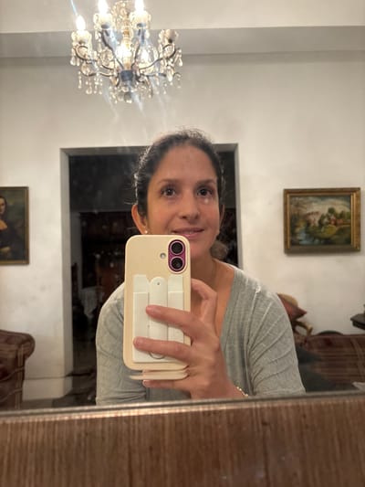 A middle-aged woman is taking a selfie in front of a mirror. The location appears to be indoors, likely a home, based on the decor. Visible elements include a crystal chandelier, framed paintings on the wall, and furniture. The city of Miraflores, Peru is not identifiable based on the visual information.