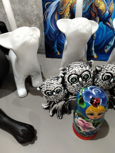 The image depicts a collection of decorative objects arranged on a flat, neutral-colored surface, likely a shelf or table. The main subjects are several ceramic figurines and a nesting doll.

In the foreground, to the left, is a dark, matte black, stylized cat's paw. To its right, a cluster of three white ceramic owl figurines with intricate black markings and bright green eyes are the focal point. The largest owl is in the center, with two smaller owls flanking it.

Next to the owls, to the right, is a vibrant blue nesting doll. The doll is hand-painted with a smiling blonde woman's face, outlined with black and adorned with rosy cheeks. She has striking blue eyes and a red bow in her hair. The doll is decorated with large pink and white flowers, green leaves, and a small, red ladybug perched on the woman's forehead.

Behind the owl figurines, two white ceramic cat figurines stand upright, facing slightly away from the viewer. They have stylized, minimalist features with pointed ears and a cross-like detail on their faces.

In the background, a brightly colored, abstract artwork is visible. It features swirling patterns in shades of deep blue, yellow, and orange, with the suggestion of a horse or mythological creature with flowing mane.

The overall impression is a curated display of artisanal decorative items. The lighting appears to be artificial, casting soft shadows and highlighting the textures of the objects. There is no visible text in the image. The setting appears to be indoors, possibly a home or a shop display.