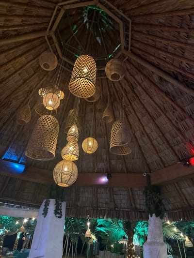The image shows a thatched-roof gazebo with numerous hanging wicker lamps. The gazebo appears to be an outdoor seating area, possibly at a restaurant or bar, with palm trees and string lights visible in the background. The lamps vary in size and shape, with some being spherical, others cylindrical, and some with intricate woven patterns. The thatched roof is a prominent feature, with straw stalks layered densely to form a protective covering. The lighting is dim, with the lamps providing warm illumination. The overall atmosphere suggests a tropical or bohemian-inspired setting. The specific location in Nesebar, Bulgaria, is not discernible from the visual information provided in the image itself.