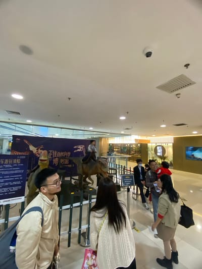 The image captures an indoor scene within a large, brightly lit commercial or exhibition space, likely Zhengjia Plaza (Grandview Mall) in Guangzhou City, China, as suggested by the visible signage. The perspective is tilted, showing a high white ceiling on the left and a busy exhibition area on the right.

In the immediate foreground, a man wearing glasses and a light beige jacket stands, looking towards the right with a neutral, observant expression. Behind him and to his left, a prominent dark blue banner showcases dinosaur imagery and Chinese text. The most discernible text reads "正佳自然科学博" (Zhengjia Natural Science Expo/Museum), indicating a natural science exhibition. Below this, smaller characters include "车厢玩须知" (Riding instructions/rules).

Central to the exhibit are two realistic dinosaur models, resembling raptors. One model appears to have a person, likely a child, sitting on its back, suggesting an interactive ride or photo opportunity. A woman with long, dark hair, wearing a light-colored sweater, stands with her back to the camera, observing these dinosaur models.

Further in the background, several more people are gathered, some appearing to be queuing behind stanchions and ropes. A person wearing a red hoodie is visible among this group. The wider setting reveals a multi-level structure with glass railings, escalators, and illuminated shopfronts, consistent with a modern shopping mall.

The ceiling is adorned with numerous recessed light fixtures, providing ample illumination, along with air vents and two security cameras. The overall atmosphere suggests an engaging and educational event, with visitors actively interacting with the dinosaur-themed natural science exhibit. The scene appears to be taking place during the daytime or during well-lit operational hours, as there are no indications of evening or night.
