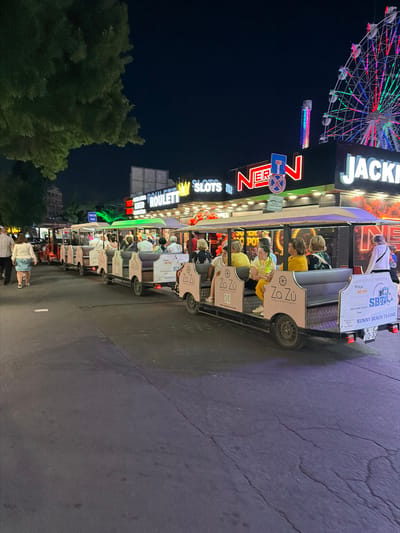 The image shows a street scene at night in the resort town of Sunny Beach, located in Bulgaria, not Nesebar. A tourist train with several open-air carriages is proceeding down the street, filled with passengers. The train has "Za Zu Lounge & Restaurant" branding on its side. To the right of the train, there is a building with neon signs that read "JACKPOT", "SLOTS", and "NERO". A Ferris wheel with colorful lights is visible in the background, as is a tall, illuminated amusement ride. People are walking on the sidewalk to the left of the train. The street is paved with asphalt and is dimly lit by the neon signs and the lights from the amusement attractions. The overall atmosphere suggests a lively evening at an amusement area.