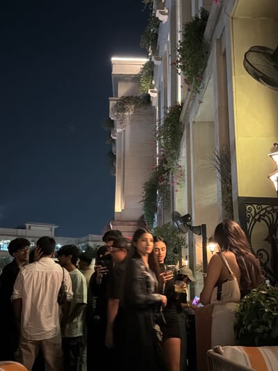 The image depicts a nighttime social gathering at an upscale outdoor restaurant or lounge within a commercial or entertainment district in New Delhi, India.
The background features a multi-story building with a light-colored facade, characterized by repeating columns, ornate balconies, and decorative ledges illuminated by strip lighting. Lush green foliage and pink flowering plants cascade from these balconies, adding natural elements to the urban structure. The sky is dark blue, indicating evening hours. To the far left, another building with a flat roofline is partially visible against the night sky. Several wall-mounted fans, dark in color, are visible on the building's facade, positioned to provide air circulation. On the far right, a section of an intricate dark metal railing or gate with scrollwork patterns is visible, beside which hangs a decorative, star-patterned lantern emitting warm light.
In the midground and foreground, approximately ten individuals are congregated, primarily engaged in conversation. On the left, a cluster of men stands, some holding drinks. One man in a white shirt and light-colored trousers, seen from the back, is central to this group, while another man with glasses smiles towards a third individual. In the center-right, two women stand prominently. One woman with long dark hair, wearing a black outfit, looks towards the left. Beside her, another woman with long dark hair holds a smartphone in her left hand and a drink in her right. Behind them, a person wearing a light-colored baseball cap holds a drink. Further to the right, a woman with long brown hair, seen from behind, wears a light-colored, possibly off-white, dress or top and carries a matching shoulder bag. She is positioned at a dark stand, appearing to interact with a tablet or screen mounted on it. In the bottom right corner, a section of an orange and white striped upholstered chair or bench is visible. The scene is illuminated by warm artificial lighting, creating a lively atmosphere.