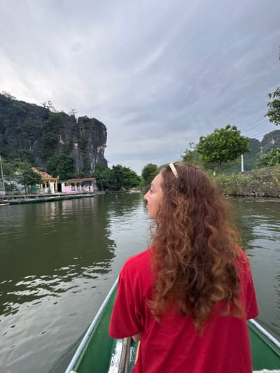 A woman with long, curly brown hair, wearing a red shirt and a gold headband, is seated in a green boat on a calm, dark body of water. She is facing to the left, looking away from the camera. In the background, a large, rugged cliff face covered in green vegetation dominates the left side of the image. At the base of the cliff, there is a light yellow building with a red-tiled roof and a pink-colored structure with a covered walkway. Several other green boats are moored along the shore. Trees and vegetation line the riverbank, and the sky is overcast with a cloudy, grey appearance. The scene appears to be in Nam Hoa Lư Ward, Vietnam, known for its karst landscapes and waterways.
