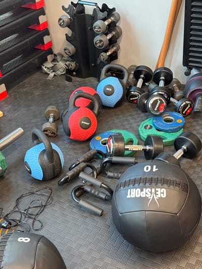 The image depicts a gym environment with various exercise equipment scattered on a black rubber mat. In the foreground, a black medicine ball with white markings, including "10" and "CEYSPORT," is prominently displayed. To its left, a black jump rope lies coiled on the mat. Also in the foreground are a pair of black push-up handles.

In the mid-ground, there are several kettlebells, one blue and one red, alongside multiple dumbbells of varying sizes. Two blue weight plates and two green weight plates are also visible. Further back, a dumbbell rack holds several dumbbells, and a long wooden pole leans against a white wall.

To the left of the frame, a set of black and red aerobic steps is stacked against the wall. The environment appears to be an indoor fitness facility. There are no people present in the image. The lighting suggests an indoor setting, possibly a commercial gym or a home gym. The image is taken from a slightly elevated angle, looking down on the equipment. The city of Kâğıthane, Turkey, is not depicted or referenced in the image itself; this information is external to the visual content.