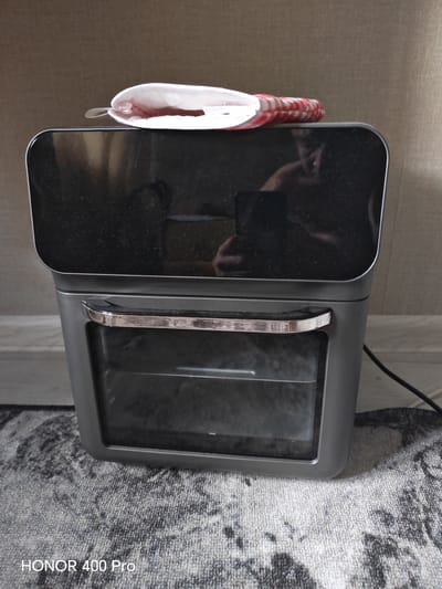 An air fryer oven is positioned on a patterned carpet in front of a wall. The air fryer is dark gray with a large black glass top that reflects a person. A chrome handle is visible on the front of the air fryer, above a glass door revealing the interior. Resting on the top of the air fryer is a white and red striped object that appears to be a fabric restraint or strap. A black power cord is visible to the right of the air fryer. The image has a watermark at the bottom left corner that reads "HONOR 400 Pro".