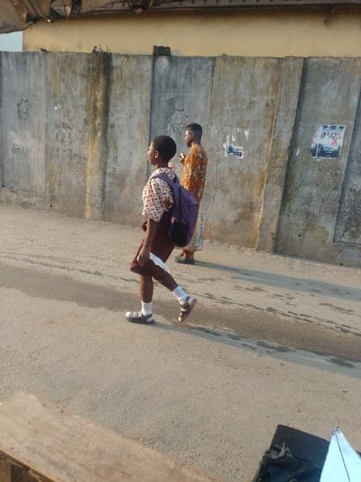 A young person in a school uniform and backpack walks on a road. The person is wearing a patterned shirt, a maroon skirt or shorts, white socks, and sandals. They are carrying something white in their hand. An adult in a patterned shirt walks behind them. They are walking alongside a tall, gray concrete wall, with a few posters attached to it. The scene appears to be outdoors during the daytime.