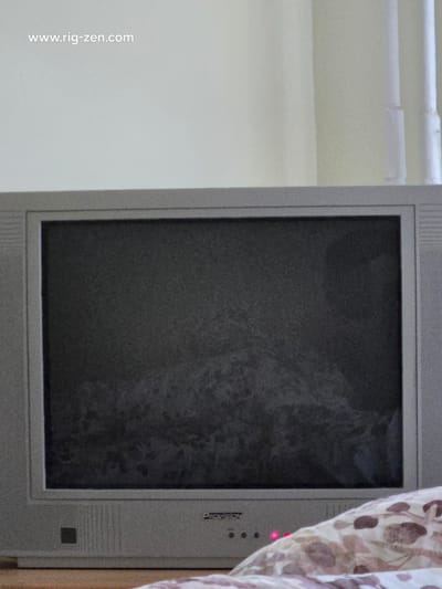 The image is a close-up, slightly blurry, eye-level shot of a vintage television set in a room. The TV is a light gray CRT model with a dark screen. The screen displays a mottled, static-like pattern. Below the screen, on the TV's frame, there is a logo that reads "VISION" and a series of small circular buttons, one of which is illuminated with a red light. The TV is placed on a surface that appears to be a bed with a patterned duvet. In the background, a light-colored wall is visible, with a section of a white cylindrical object, possibly a pipe or radiator cover. The top left corner of the image contains a watermark that reads "www.rig-zen.com". The image does not provide any visual information that would indicate its location in Jõhvi, Estonia.