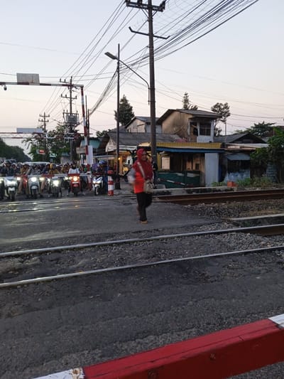 The image shows a street scene in Semarang, Indonesia at a railroad crossing. A woman wearing an orange top and headscarf is crossing the railroad tracks. A line of motorbikes is stopped behind a railroad barrier. Power lines are visible overhead, with buildings in the background. The setting suggests an urban area in Semarang.