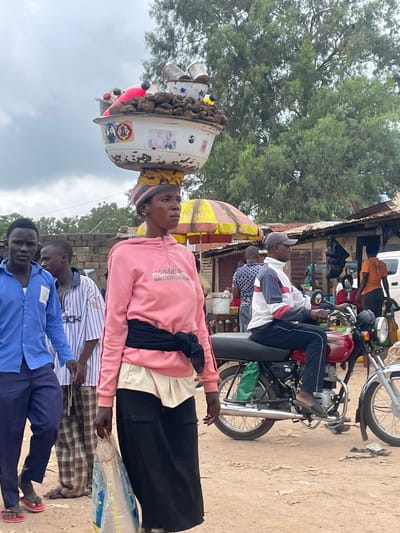 The image depicts a street scene in Wang, Nigeria, with a woman carrying a large bowl of goods on her head. She is wearing a pink hooded sweatshirt and a black belt. In front of her, a man in a blue shirt and dark pants walks with two other men. To the right, a man in a white, blue, and red jacket is sitting on a red motorcycle. Several market stalls and trees are visible in the background, suggesting a bustling environment. The ground is dusty, typical of a dry, outdoor market setting.