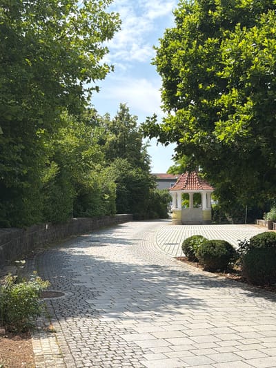 The image shows a paved pathway curving through a park-like setting. Large green trees line the pathway, casting shadows on the light-colored stone pavement. On the right side of the path, there are several rounded bushes. In the distance, a small, open-sided structure with a red tile roof supported by white columns is visible. The sky above is blue with some scattered white clouds. The pathway appears to be made of cobblestones and rectangular pavers. There are no people visible in the image. The scene suggests a public outdoor space in Ebermannstadt, Germany, likely a park or a garden area.