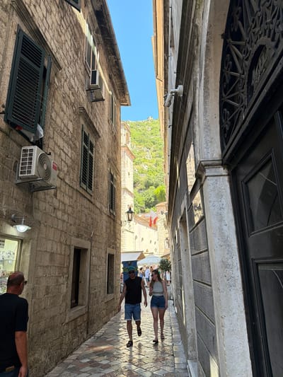 In Kotor, Montenegro, a narrow cobblestone street is lined with old stone buildings. Sunlight streams down, creating sharp shadows and illuminating the worn textures of the architecture. On the left, a building features green shutters, an air conditioning unit, and a modern light fixture. On the right, a building has a dark, ornate door and a decorative metal grate.

Two individuals are walking away from the camera down the street. The person in front, on the right, is a man wearing a black sleeveless shirt, denim shorts, a green baseball cap, and sandals. The person behind him, on the left, is a woman wearing a light-colored tank top and denim shorts, with sandals. They are walking in the same direction.

Further down the street, other people can be seen browsing shops and walking. In the background, a steep, verdant hillside rises, crowned with clear blue sky. A white and red flag flies from a building in the distance. The overall scene depicts a vibrant street in a historic European city.