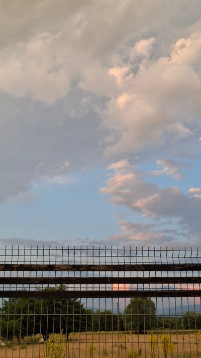 The image depicts a natural scene with a fence in the foreground. The fence is made of metal mesh with vertical wooden posts, suggesting it might be a boundary or part of a structure. Beyond the fence, a landscape unfolds with trees and vegetation. The sky is filled with clouds, tinged with pink and orange hues, indicating that it is either sunrise or sunset. The lighting suggests a soft, warm glow, typical of these times of day. The presence of trees and what appears to be a field or meadow suggests a rural or semi-rural location. The image does not contain any people or visible text. The overall mood is tranquil and serene, capturing the beauty of the sky during twilight. The orientation of the image is portrait.