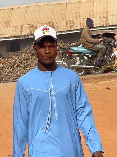 The image depicts an outdoor scene in Zaria, Nigeria. In the foreground, a young Black male is centered, facing the camera with a neutral expression. He wears a white and black baseball cap with multi-colored stylized text "AKO BIG" on the front, and a light blue long-sleeved traditional tunic featuring white piping details and a decorative tassel at the neckline. A small dark earpiece is visible in his right ear. The ground beneath him is reddish-brown dirt.
In the mid-ground, slightly to the left, is a large pile of light-colored dry wooden branches or firewood. To the right of this pile, a second Black male is seated on or operating a black motorcycle with a light blue seat, facing right, away from the camera. This individual wears a brown jacket, dark pants, a dark blue beanie, and a dark face covering. A second white motorcycle is partially visible behind the black one.
The background features a building with a light brown corrugated metal roof and walls, positioned behind the woodpile. The sky above is clear and light blue, indicating bright daylight conditions.