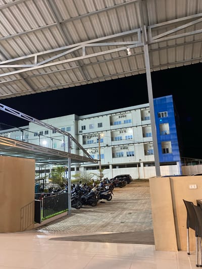 A nighttime scene in Banda Aceh, Indonesia, features a multi-story building with a blue facade on its upper section. The building has numerous windows, and some appear to be lit from within. A metal roof structure with corrugated panels is visible in the foreground, supported by a metal frame. Adjacent to the building is a paved area with a brick pattern, where several motorcycles are parked in a row. The lighting suggests it is evening or night. The overall setting appears to be an urban or semi-urban environment, possibly a commercial or residential complex. There is no visible text in the image.