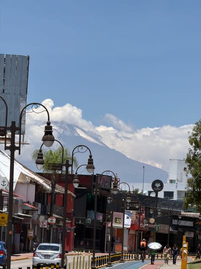 The image shows a street scene in San Andrés Cholula, Mexico, featuring a partial view of Popocatépetl volcano shrouded in clouds. The foreground includes streetlights, signage, buildings with thatched roofs, a variety of commercial storefronts, and pedestrians. A taxi is visible on the street.